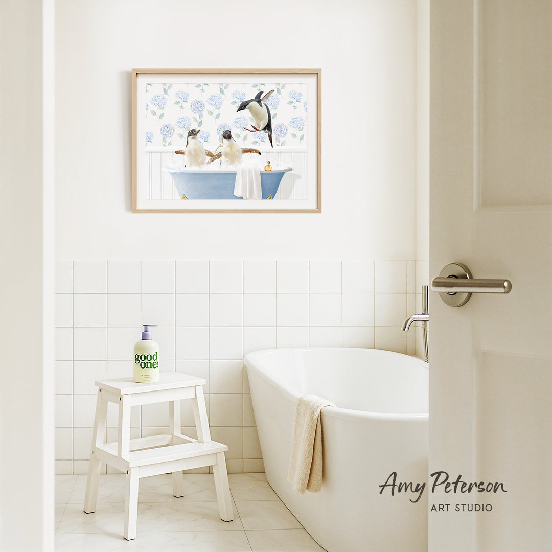 A framed painting of penguins in a bathtub hangs on a white tiled wall above a white bathtub in a bathroom.