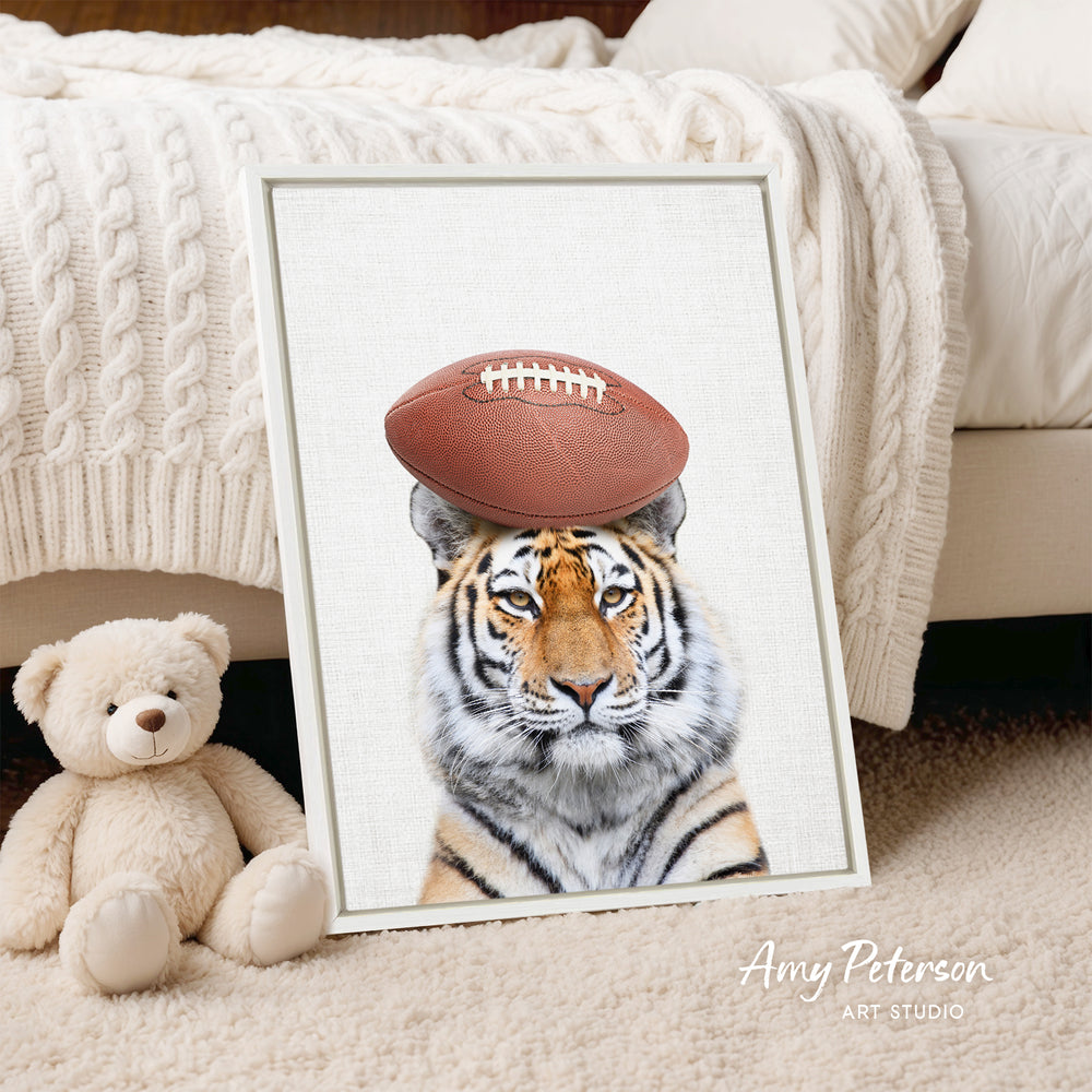 A framed print of a tiger wearing a football on its head is displayed on a bed, with a teddy bear next to it.