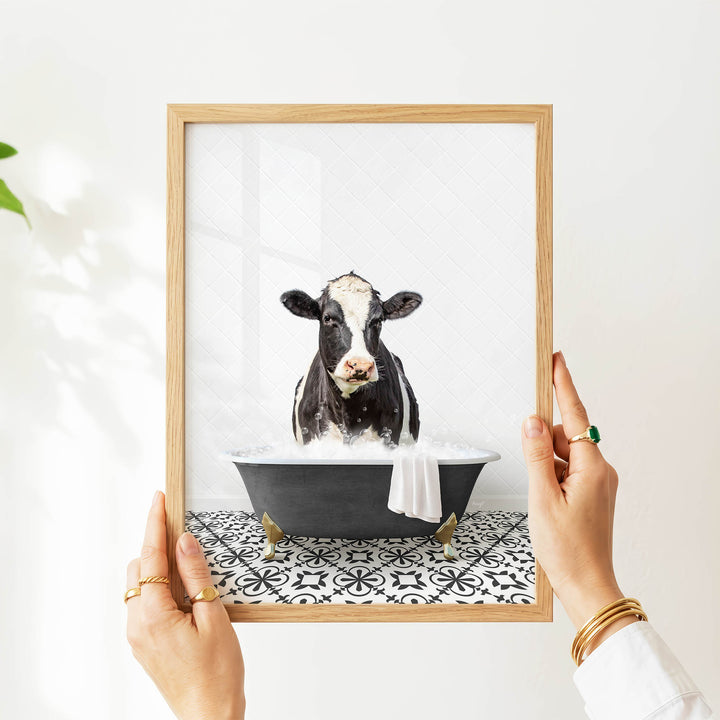 A person's hands are holding a picture frame with a framed image of a cow sitting in a bathtub filled with water.