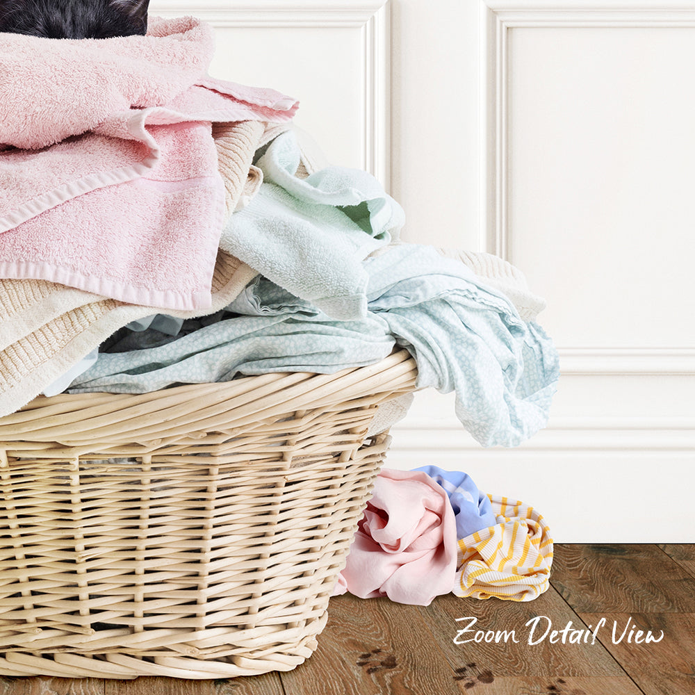A wicker laundry basket filled with various folded clothes, including a pink towel, sits on a wooden floor against a white wall.