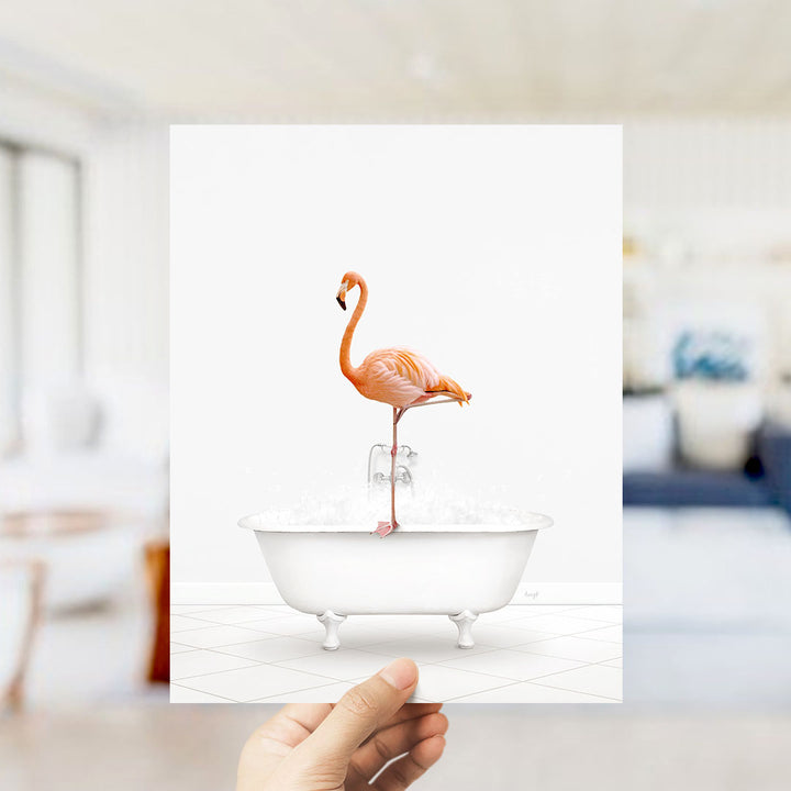 A hand holds up a framed image of a flamingo standing in a bathtub filled with bubbles.
