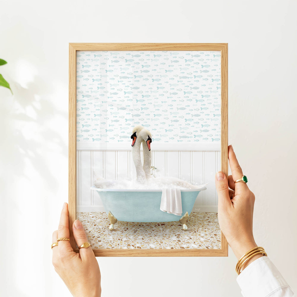 A person's hands are holding a framed picture of a swan sitting in a bathtub filled with bubbles, with a patterned wall behind it.