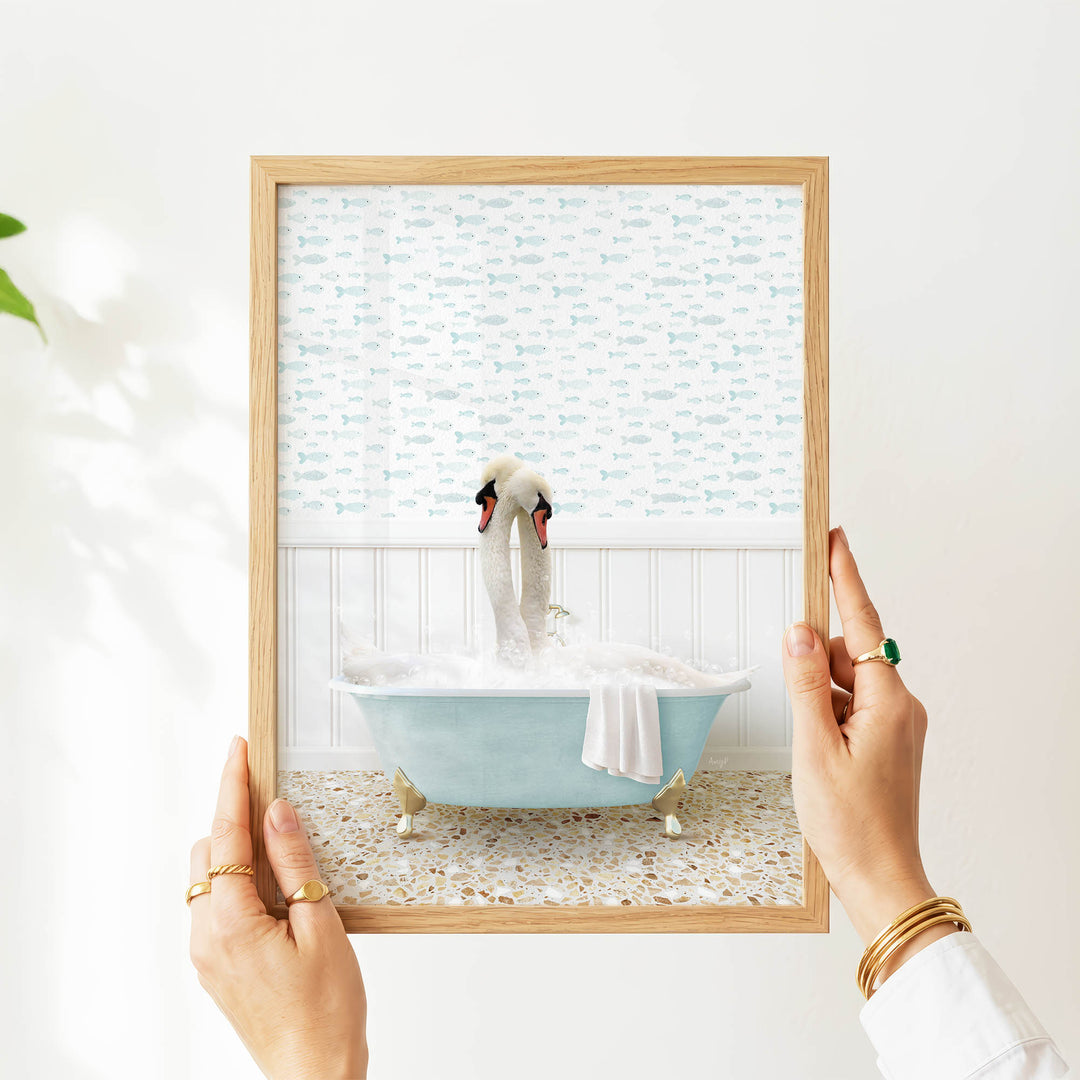 A person's hands are holding a framed picture of a swan sitting in a bathtub filled with bubbles, with a patterned wall behind it.