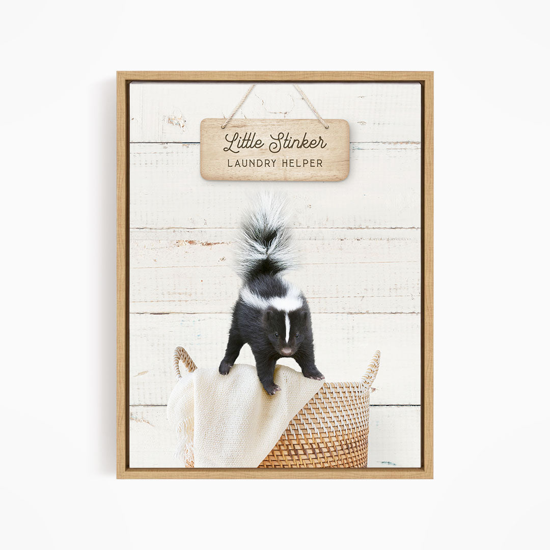 A framed image of a black and white skunk standing on a woven basket, with a wooden sign above it that reads "Little Stinker Laundry Helper".