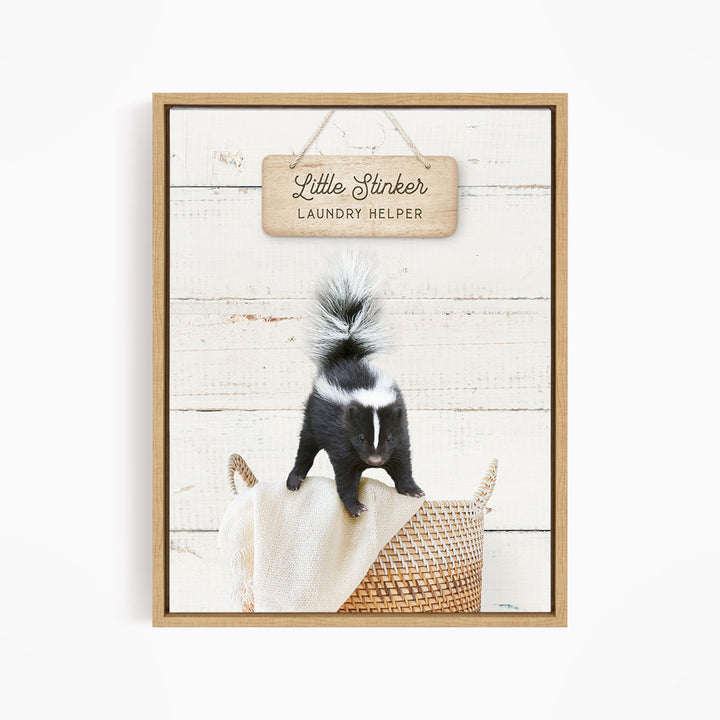 A framed image of a black and white skunk standing on a woven basket, with a wooden sign above it that reads "Little Stinker Laundry Helper".