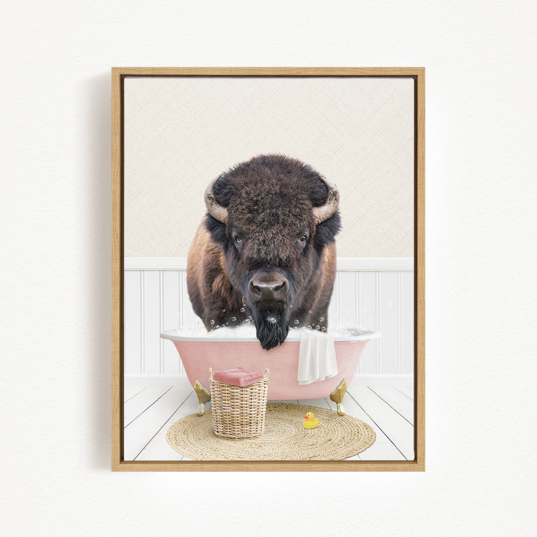 A buffalo is sitting in a pink bathtub filled with water, surrounded by a basket of toys and a yellow rubber duck.
