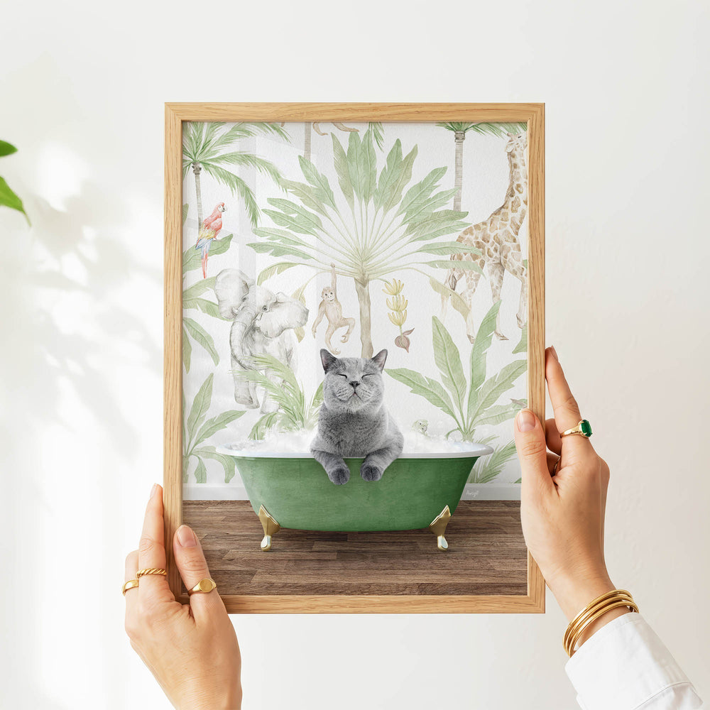 A person's hands are holding a framed picture of a cat sitting in a green bathtub, surrounded by tropical plants and animals.