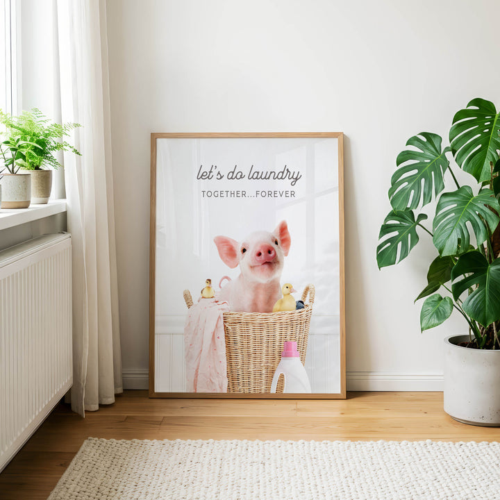 A framed poster with a pig sitting in a basket, surrounded by various objects, is displayed on a wall in a room with a rug and potted plants.