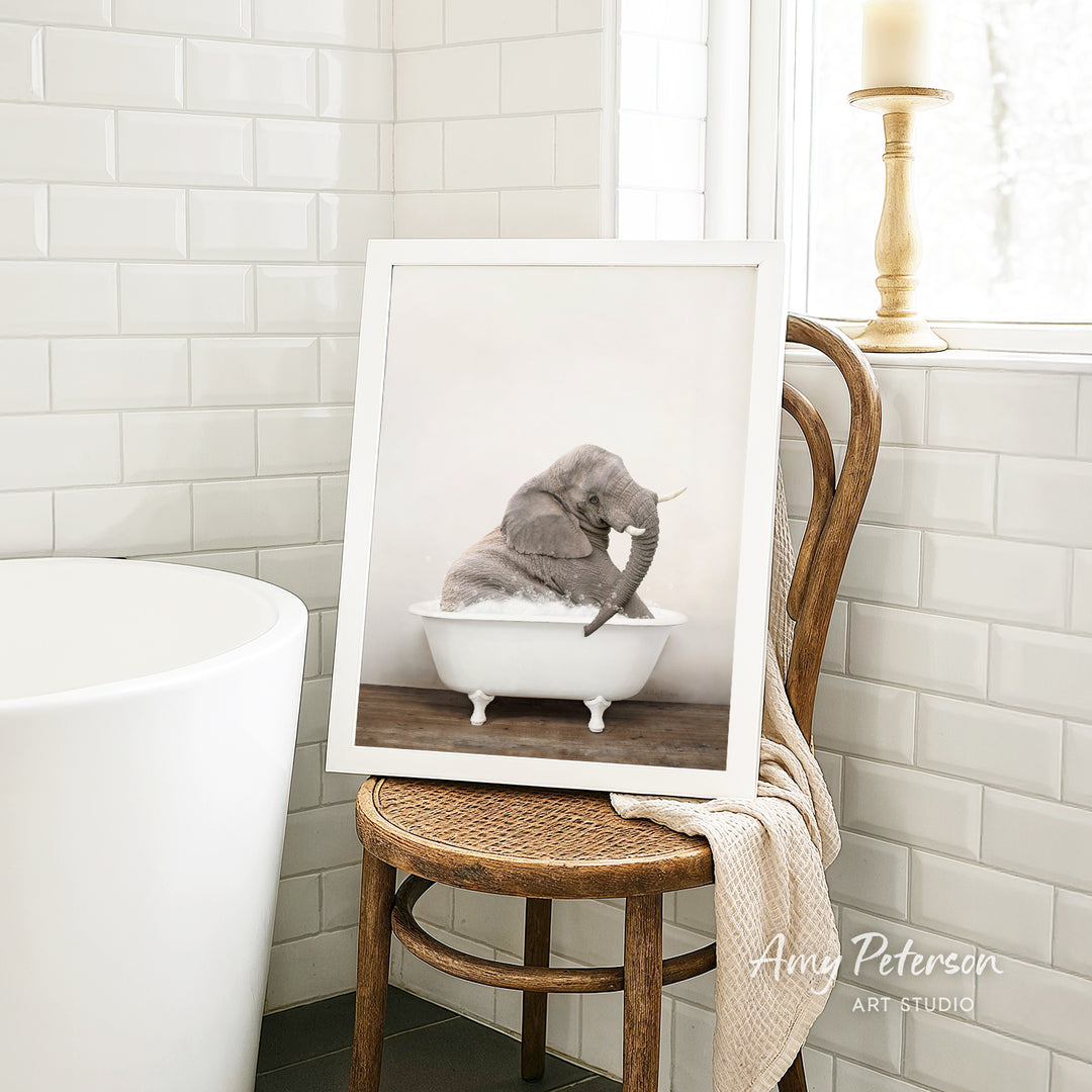 A framed photograph of a baby elephant sitting in a bathtub is displayed on a wooden chair in a bathroom setting.