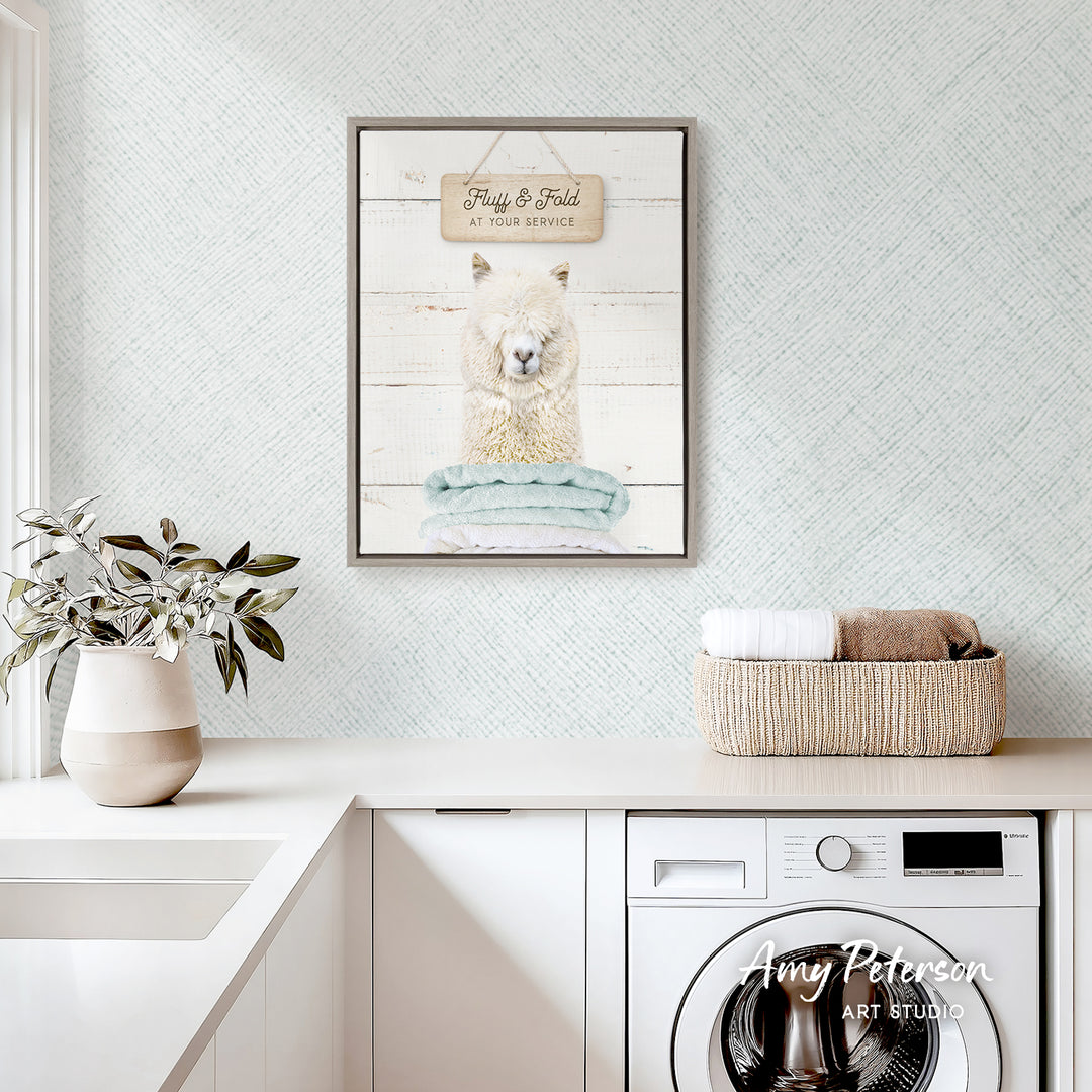 a framed print of a llama on a wall above a laundry basket, with a potted plant and a white sink visible in the foreground.