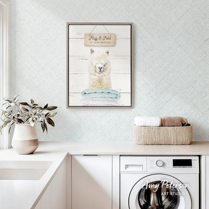 a framed print of a llama on a wall above a laundry basket, with a potted plant and a white sink visible in the foreground.