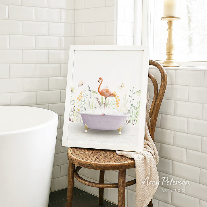 A framed painting of a pink flamingo sitting in a purple bathtub, surrounded by flowers and plants, is displayed on a wooden chair in a bathroom setting.