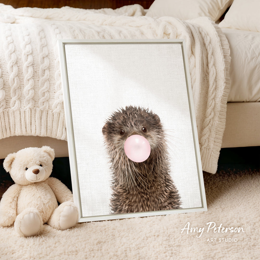 A framed image of an otter blowing a bubble with a pink bubble gum bubble, accompanied by a teddy bear on a bed.