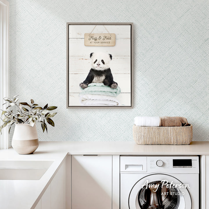 a framed picture of a panda bear sitting on a stack of towels, with a sign above it that says "Please & Fold at Your Service". The panda bear is sitting on a stack of white towels, and there is a basket of towels next to the panda bear. The scene is set in a room with a white countertop and a white washing machine.