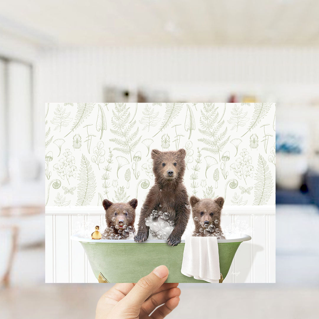 A hand holds up a framed image depicting three adorable brown bears sitting in a green bathtub, surrounded by bubbles and a towel.