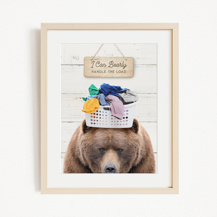 A brown bear sits atop a laundry basket filled with clothes, with a wooden sign above that reads "I Can Bearly Handle the Load".