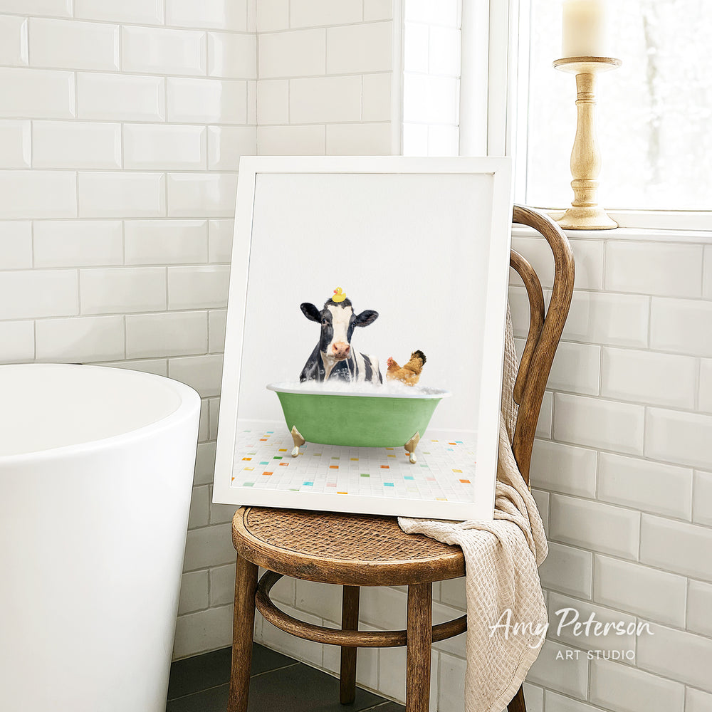 A framed image of a cow sitting in a green bathtub, with a chicken nearby, is displayed on a wooden chair in a bathroom setting.