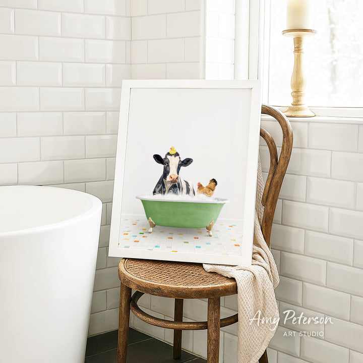 A framed image of a cow sitting in a green bathtub, with a chicken nearby, is displayed on a wooden chair in a bathroom setting.