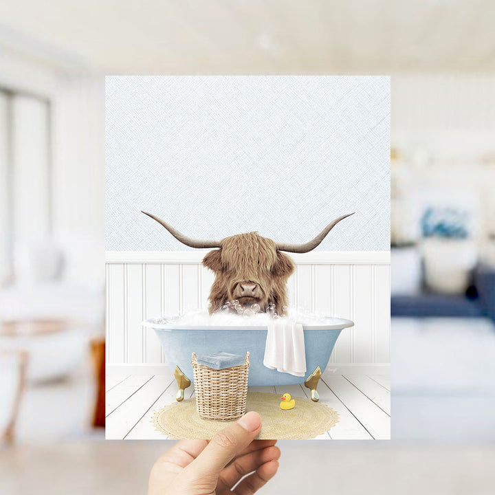 A hand holds up a framed image depicting a Highland cow with long horns sitting in a bathtub filled with bubbles, surrounded by a yellow rubber duck and a white towel.