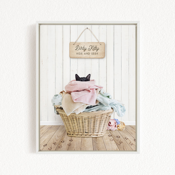 A black cat is sitting in a wicker basket filled with clothes, with a wooden sign above the basket that reads "Dirty Kitty HIDE AND SEEK".
