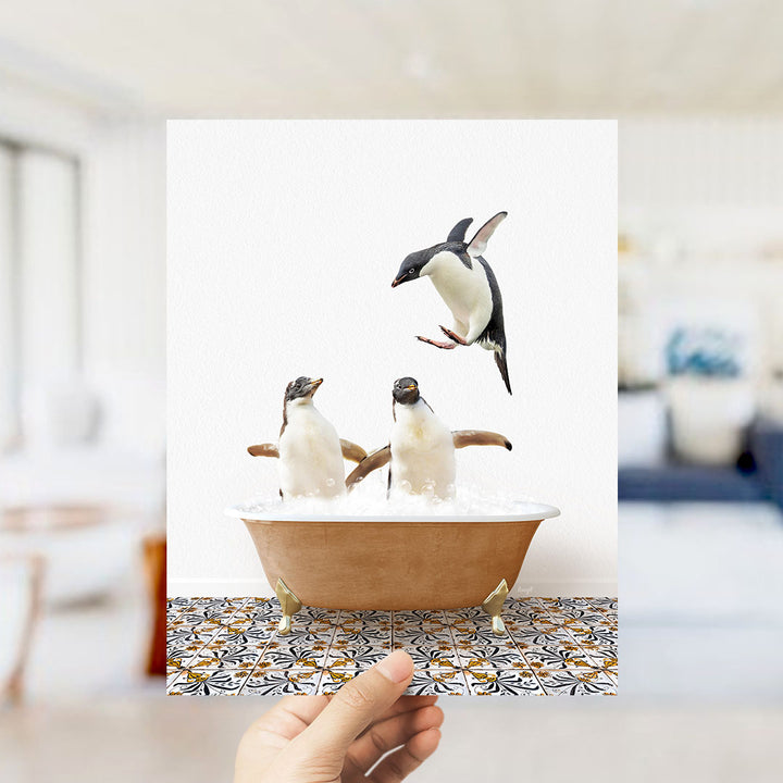 A hand holds up a framed image depicting two penguins in a bathtub, with one penguin flying out of the tub.