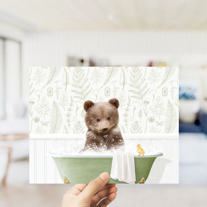 A hand holding a framed print of a cute brown bear sitting in a bathtub, surrounded by a green bathtub, a yellow duck, and a white towel.