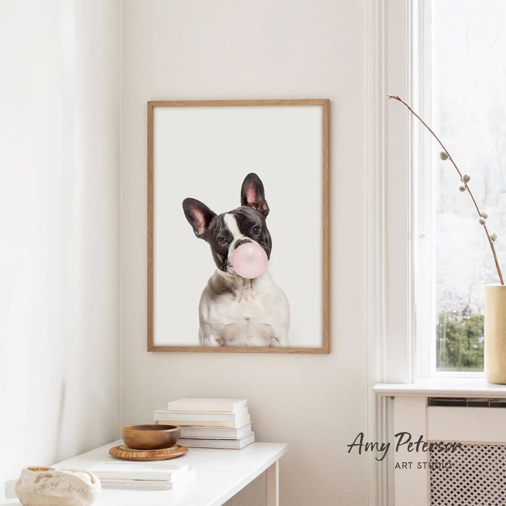 A framed image of a dog blowing a bubble gum bubble is hanging on a white wall in a room with a white table and a window.