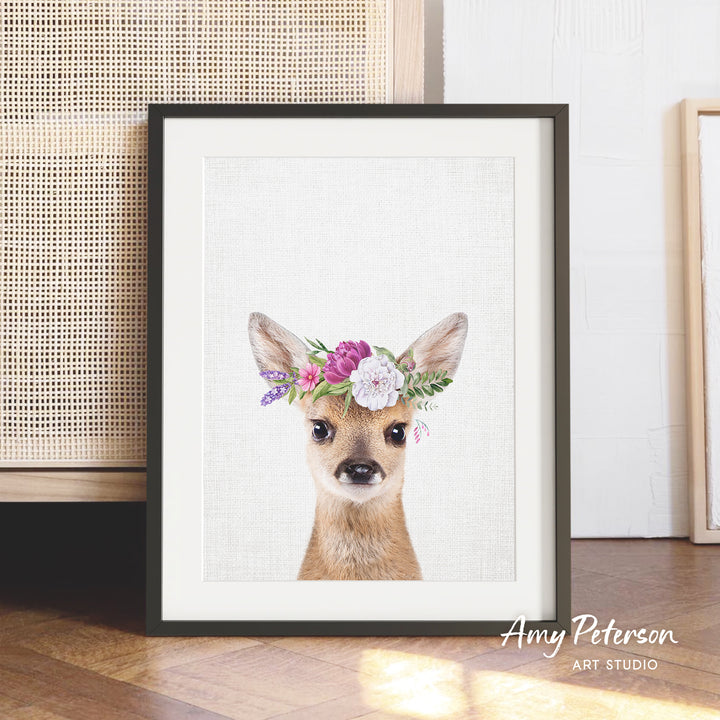 A framed print of a deer wearing a floral crown, with a white background and a wooden floor in the background.