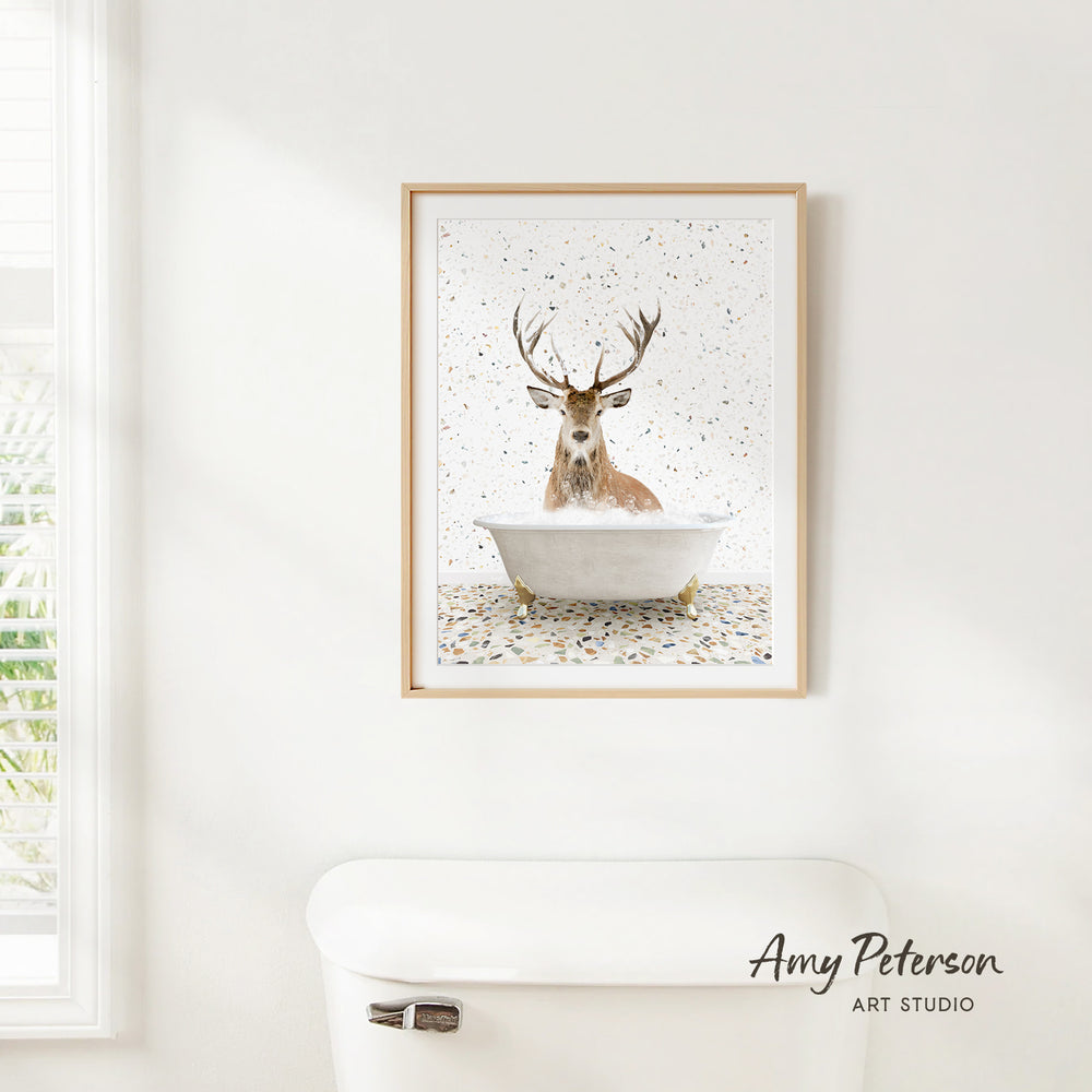 A framed print of a deer sitting in a bathtub, surrounded by confetti, hangs on a white wall.