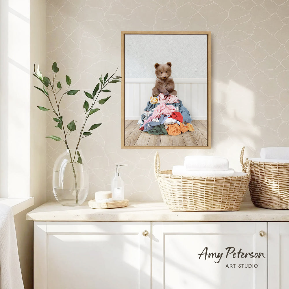 a framed photograph of a teddy bear sitting on a pile of laundry, hanging on a wall above a white cabinet.