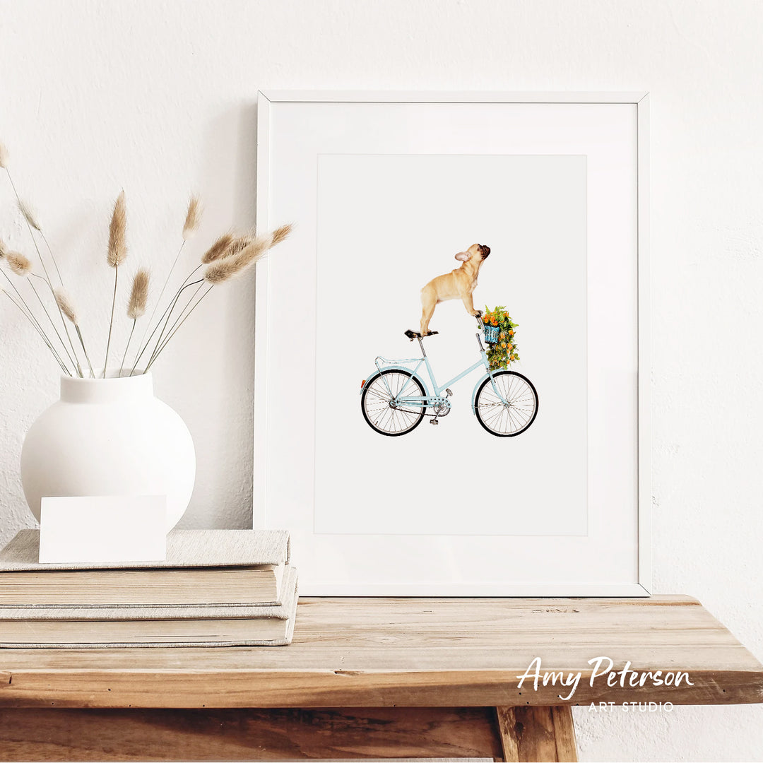 a framed illustration of a dog riding a bicycle, with a vase of dried flowers and a stack of books on a wooden shelf in the foreground.