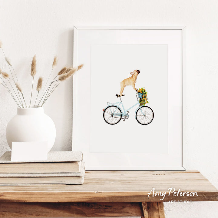 a framed illustration of a dog riding a bicycle, with a vase of dried flowers and a stack of books on a wooden shelf in the foreground.
