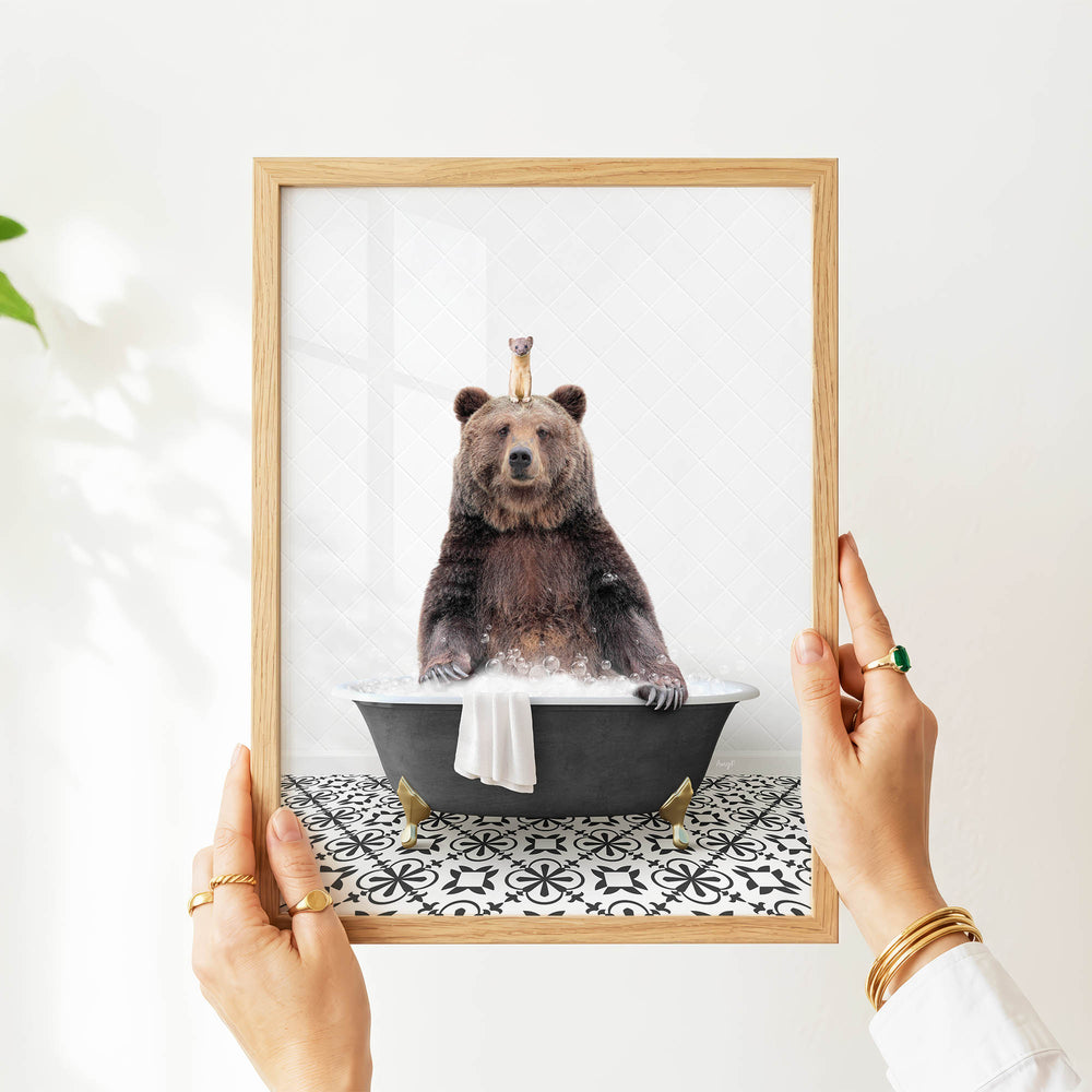 A person's hands are holding a framed picture of a bear sitting in a bathtub, with a small bird perched on its head.