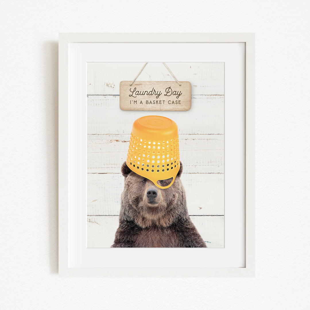 A framed image of a bear wearing a yellow laundry basket on its head, with a sign above that says "Laundry Day I'm a basket case".