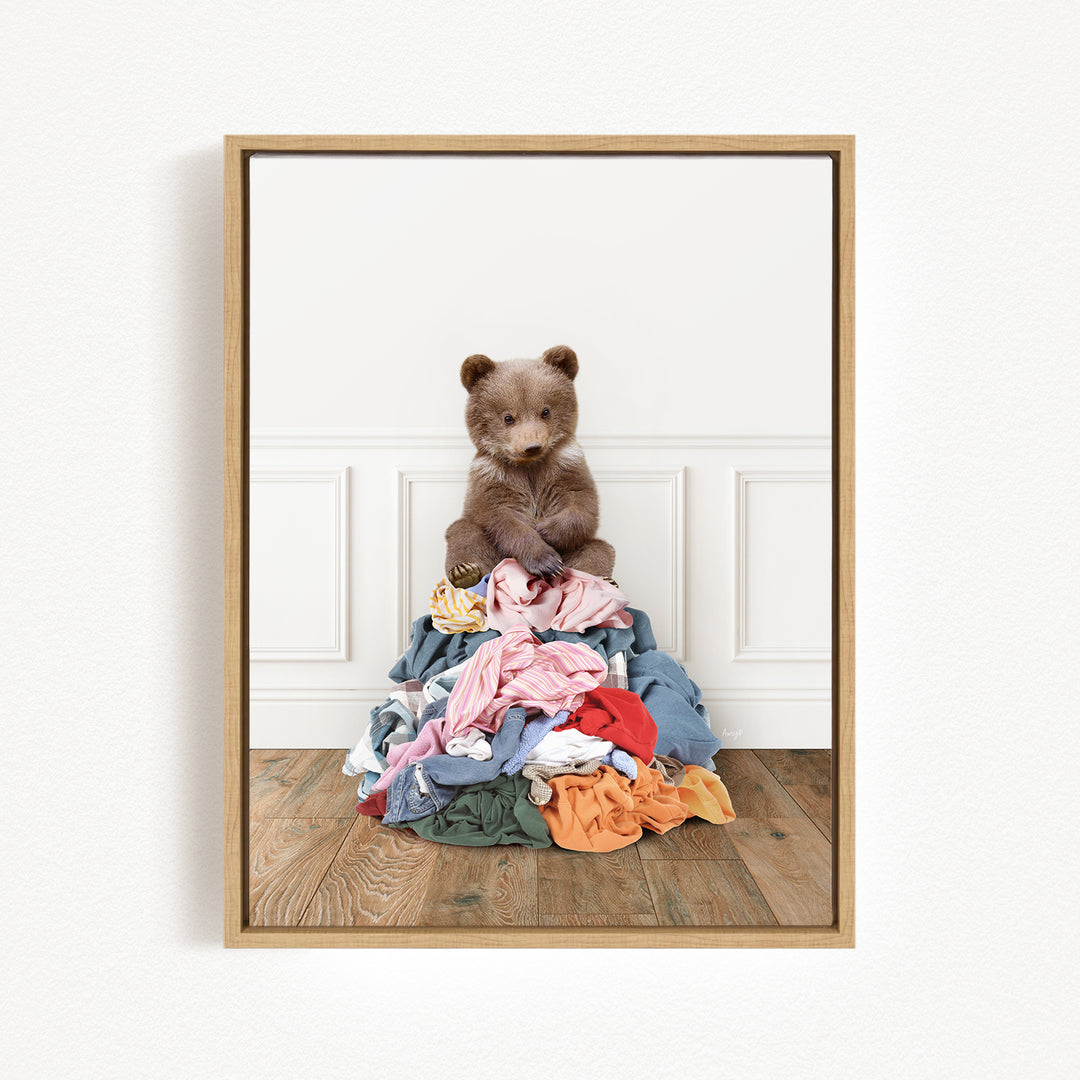 A brown bear sits atop a pile of clothes on a wooden floor, with a white wall and paneled wall in the background.