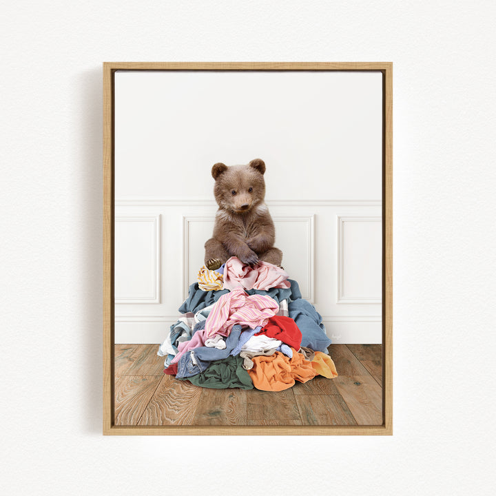 A brown bear sits atop a pile of clothes on a wooden floor, with a white wall and paneled wall in the background.