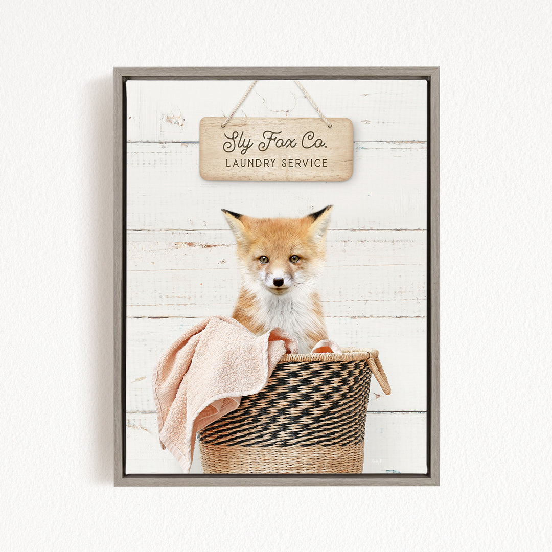 A framed image of a young fox sitting in a woven laundry basket, with a sign above that reads "Fly Fox Co. Laundry Service".