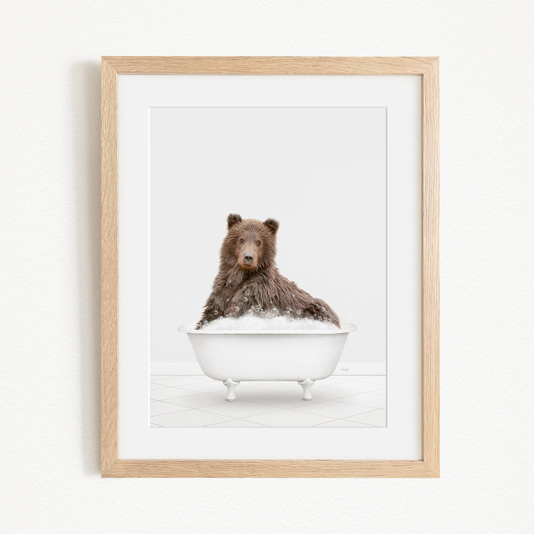A brown bear sitting in a white bathtub filled with bubbles, looking directly at the camera.