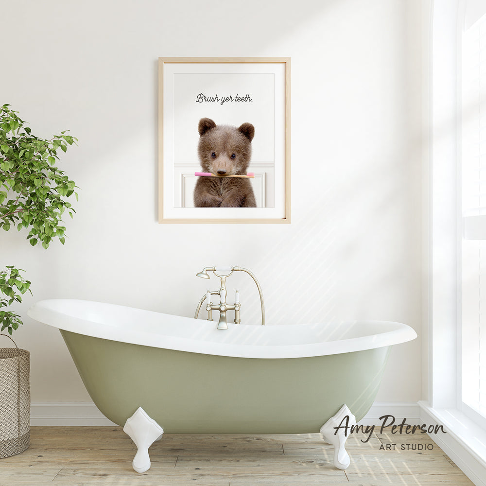 A framed image of a brown bear holding a toothbrush in its mouth is displayed on a white wall above a green clawfoot bathtub.