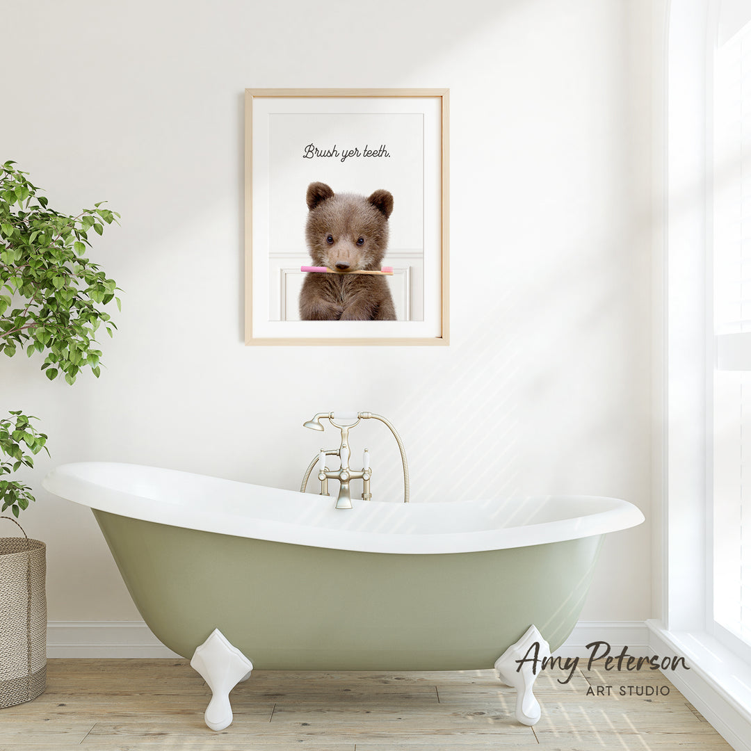 A framed image of a brown bear holding a toothbrush in its mouth is displayed on a white wall above a green clawfoot bathtub.