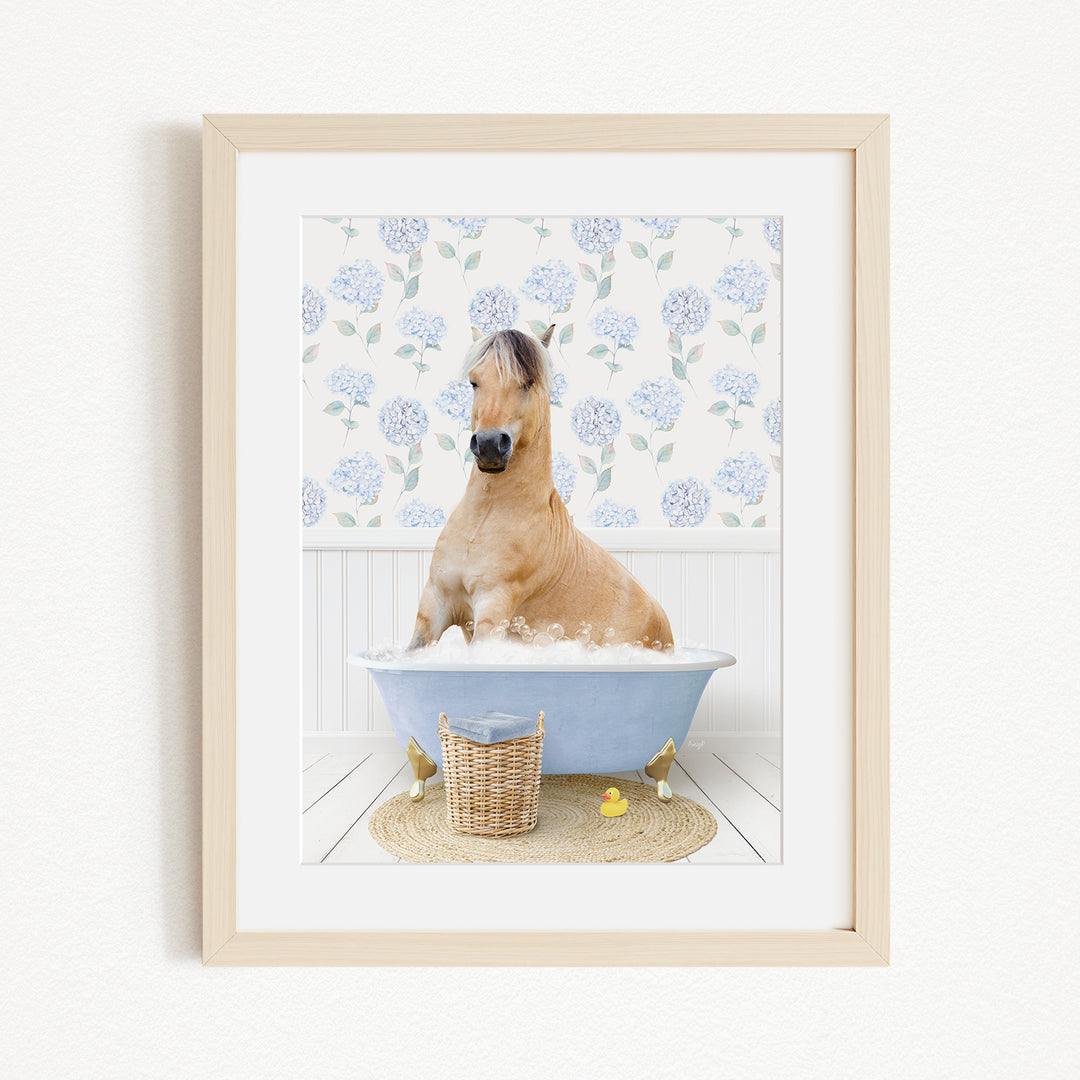 A light brown horse is sitting in a blue bathtub filled with bubbles, surrounded by a white floral wallpaper and a wicker basket.