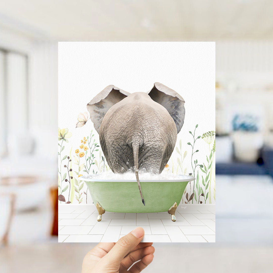 A hand holds up a framed image of an elephant sitting in a green bathtub filled with water, surrounded by flowers and plants.