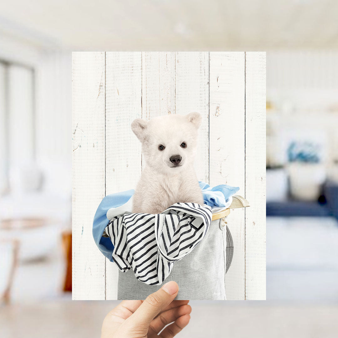 A small white polar bear cub sitting in a basket filled with clothes.