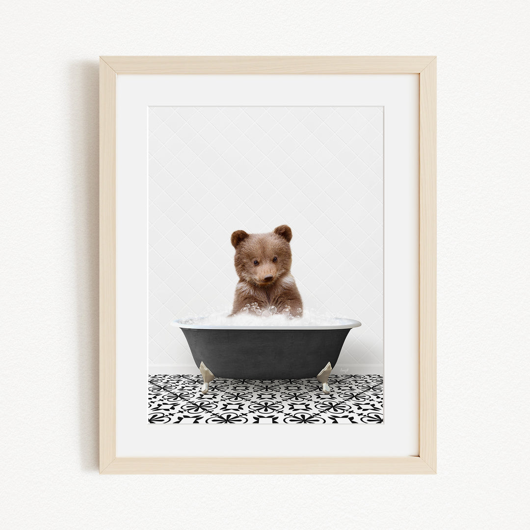 A cute brown bear cub sitting in a black bathtub filled with water, surrounded by a black and white patterned tile floor.