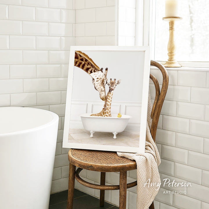 A framed image of two giraffes sitting in a bathtub, with one giraffe appearing to be bathing the other. The image is displayed on a wooden chair in a bathroom setting.