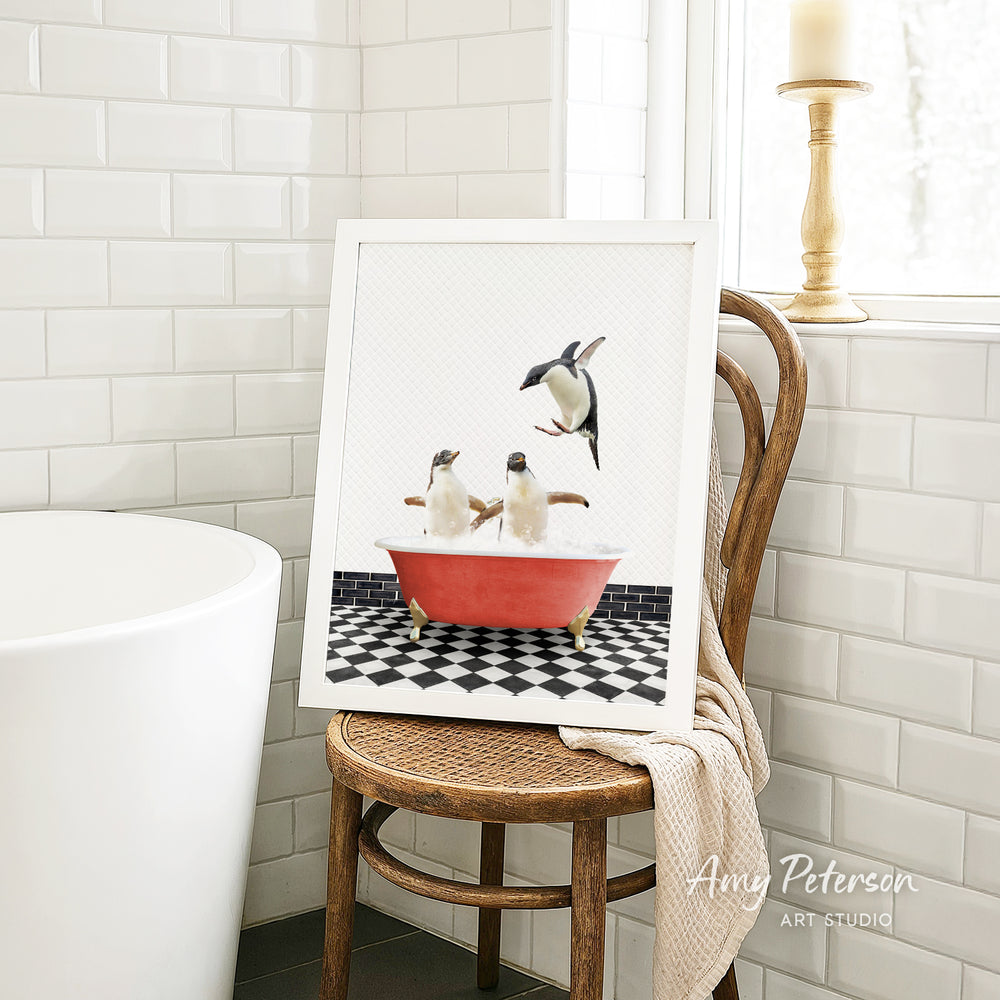 A framed image of two penguins sitting in a red bathtub, surrounded by a checkered black and white floor, is displayed on a wooden chair in a bathroom setting.