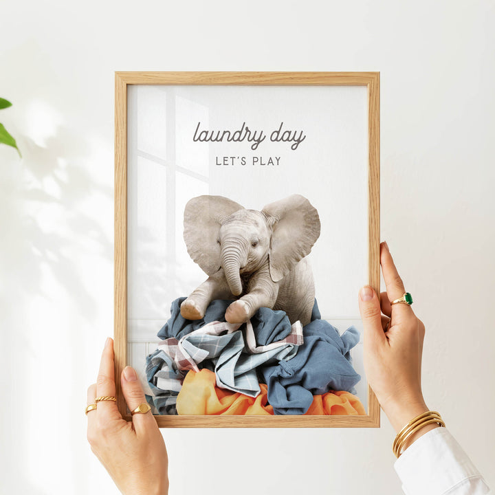 A person's hands are holding a framed picture of a baby elephant sitting on a pile of clothes, with the text "laundry day let's play" written above the image.