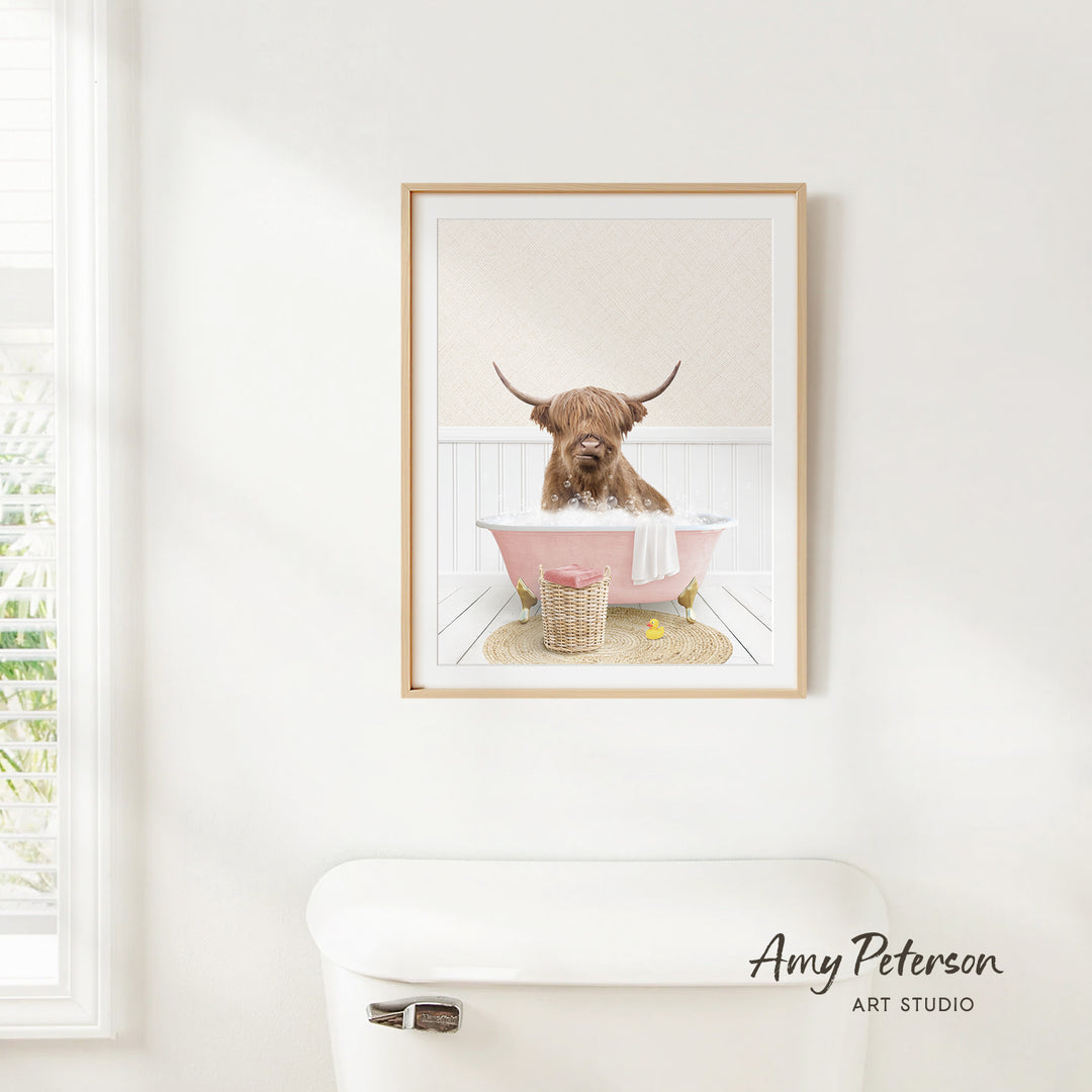 A framed image of a Highland cow sitting in a pink bathtub, with a towel and other items around it, is hanging on a white wall.