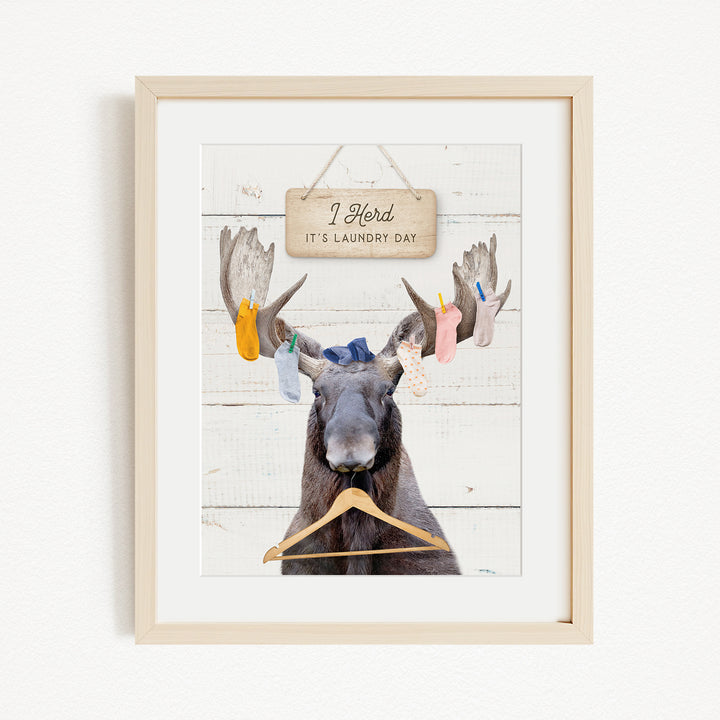 A framed image depicting a moose with a sign that says "I herd it's laundry day" hanging on a wooden background.
