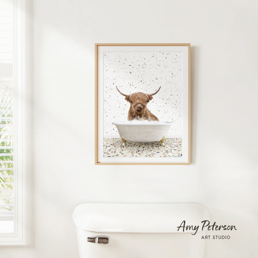 A framed image of a Highland cow sitting in a bathtub, surrounded by a speckled background, is hanging on a white wall.